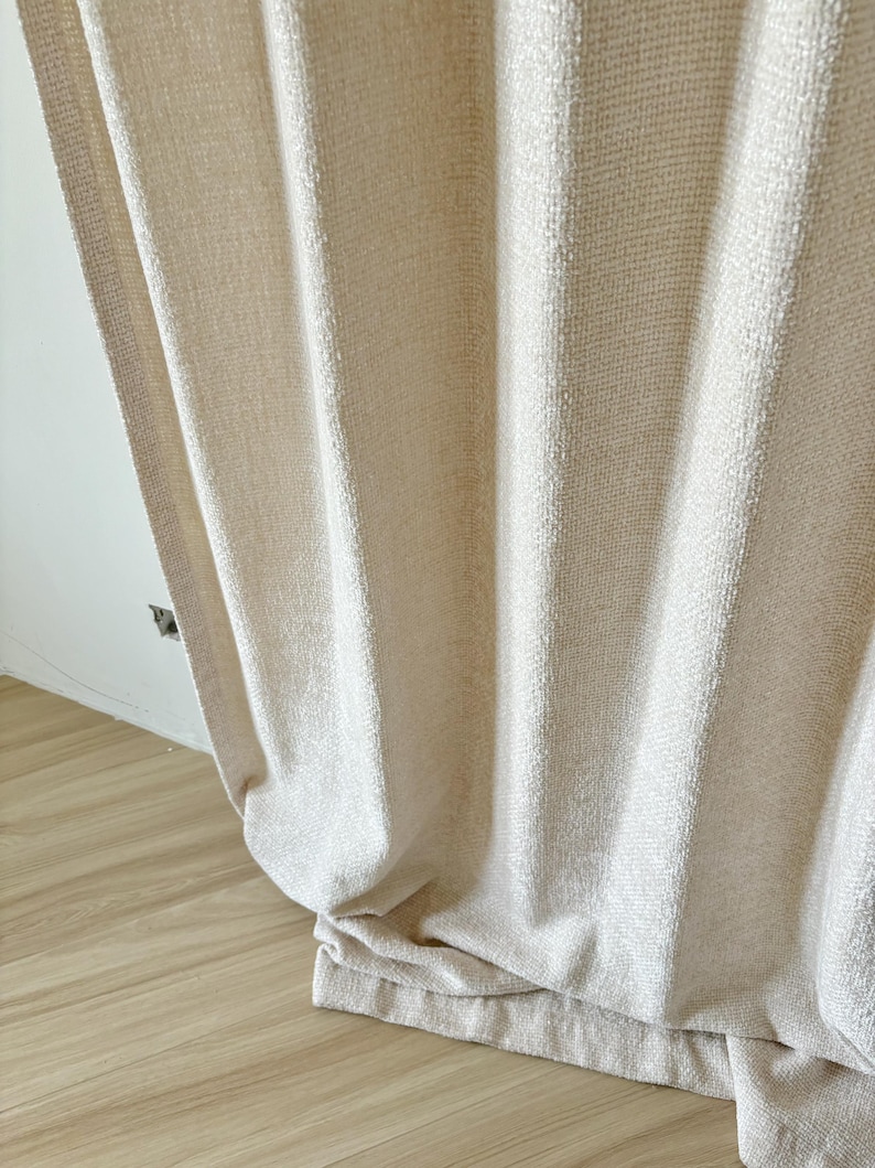 May include: A floor-length, off-white curtain featuring a textured, woven fabric with vertical stripes. The material has a subtle, slightly shimmering finish and hangs in soft, loose folds that extend down to the floor, creating a draped effect.