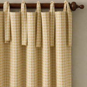 Può includere: Un pannello per tenda a quadretti vichy gialli e bianchi con un design a laccetti per il fissaggio. Il tessuto è appeso a un bastone per tende in legno marrone scuro con terminali decorativi, creando morbide pieghe verticali lungo il telo.