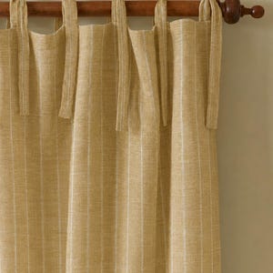 May include: Tan-colored linen curtains with vertical white stripes, hanging from a wooden rod. The curtains are tied with matching fabric ties. The rod has a decorative finial on the end.