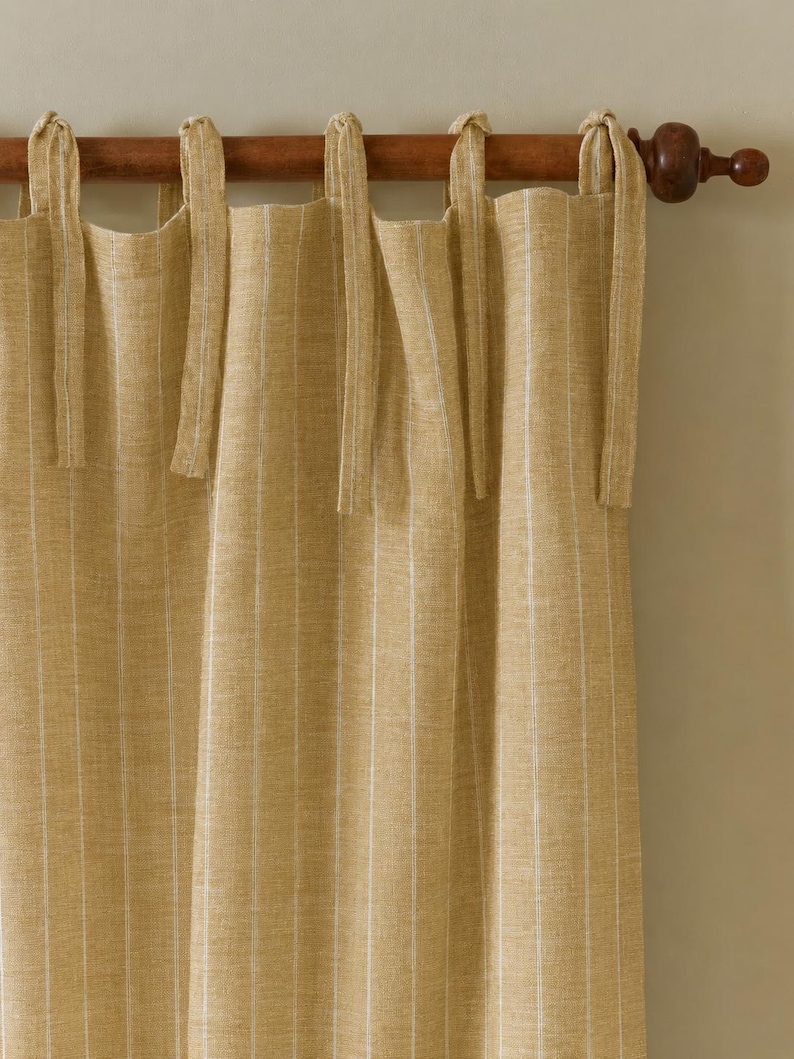 May include: Tan-colored linen curtains with vertical white stripes, hanging from a wooden rod. The curtains are tied with matching fabric ties. The rod has a decorative finial on the end.
