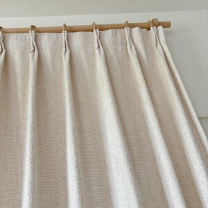 May include: A cream-colored curtain with a textured, woven fabric finish featuring vertical pinch pleats. The curtain is suspended from a light-toned wooden rod by matching wooden rings, creating uniform folds that drape downward from the top header.