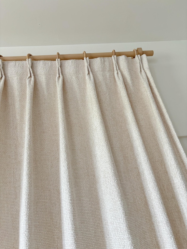 May include: A cream-colored curtain with a textured, woven fabric finish featuring vertical pinch pleats. The curtain is suspended from a light-toned wooden rod by matching wooden rings, creating uniform folds that drape downward from the top header.