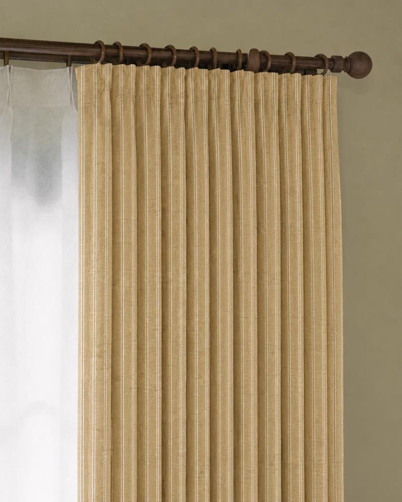 May include: Beige and white striped curtain panel hanging from a dark brown rod with decorative rings. The curtain is pleated and has a textured appearance, creating vertical lines. A sheer white curtain is visible to the left.