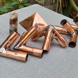 May include: A collection of polished copper pipe fittings featuring various angled joints and connectors, including L-shaped and multi-directional pieces, arranged alongside a small cylindrical resin tower containing embedded multi-colored stones.