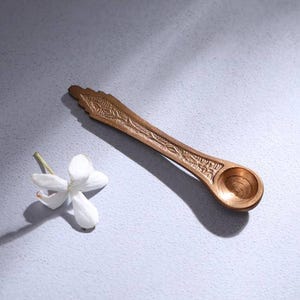 May include: A small, metallic, copper-toned spoon featuring an elongated handle with intricate, embossed linear patterns and a circular bowl. Beside the spoon lies a single, delicate white flower with four petals and a thin green stem.