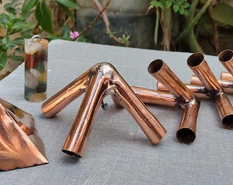 Puede incluir: Una colección de objetos de cobre que incluye una pirámide pulida, un colgante cilíndrico relleno de piedras multicolores y cuatro conectores de tubería de cobre con juntas de tres vías, dispuestos sobre una superficie plana.