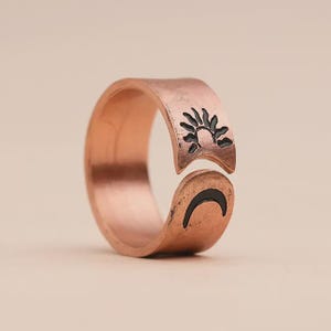 Op de afbeelding: Een open koperen ring met een gehamerde textuur en een gepolijste afwerking. De band toont aan het ene uiteinde een zwart gegraveerd zonmotief en aan het andere uiteinde een zwart gegraveerd maansikkelmotief, wat zorgt voor een hemels ontwerp.