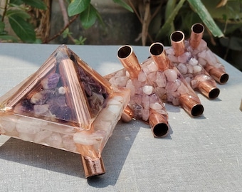 May include: A pyramid-shaped resin object containing light pink stones and copper pipes, a rectangular resin block with copper pipes and pink stones, and a cylindrical resin pendant with embedded stones and a silver-toned metal bail.