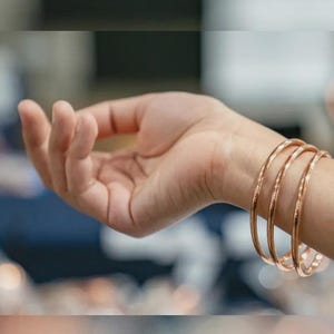 以下が含まれることがあります： 手首に装着された、ローズゴールド調の磨き上げられた金属製の細いバングル3本セット。ブレスレットは滑らかで丸みを帯びた表面と光沢のある仕上げが特徴で、3つの円形のバンドすべてにおいてシンプルで統一感のある外観を作り出しています。