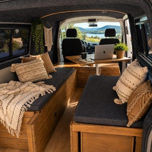 Könnte beinhalten: Das Innere eines Campingbusses zeigt zwei Holzsitzbänke mit dunkelgrauen Polstern, einen zentralen Holztisch und einen Laptop. Der Raum ist mit cremefarbenen Kissen mit Quasten, einer strukturierten Decke und einer kleinen Topfpflanze ausgestattet.