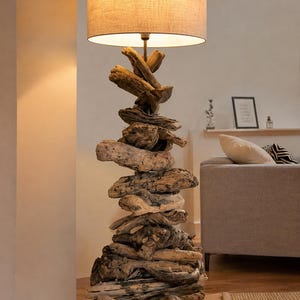 May include: A floor lamp featuring a base constructed from a vertical stack of weathered, natural driftwood pieces in varying brown and gray tones, topped with a cylindrical, light beige fabric lampshade that emits a warm, soft glow.