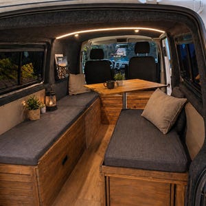 Könnte beinhalten: Innenansicht eines Campingbusses mit Holzbänken, grauen Kissen und einem kleinen Tisch. Der Bus hat eine warme, einladende Atmosphäre mit sanfter Beleuchtung und dekorativen Kissen. Die Bänke scheinen fest eingebaut zu sein.
