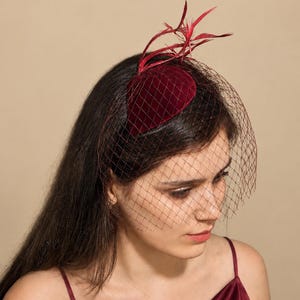 Könnte beinhalten: Ein dunkelroter Fascinator aus Samt mit einer strukturierten, abgerundeten Basis, verziert mit aufrechten roten Federn und einem passenden roten Schleier, der über die Vorderseite fällt, kombiniert mit einem dunkelroten Satin-Top.