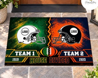 May include: A rectangular doormat featuring a split design with a white football helmet on a green background and a black football helmet on an orange background. Text includes TEAM 1, TEAM 2, 1919, 1920, and HOUSE DIVIDED with a football graphic.