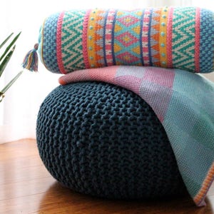 May include: A blue knitted pouf with a colorful patterned bolster pillow and a light blue and pink throw blanket on top.