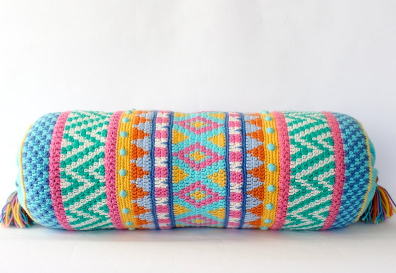 May include: A colorful crocheted bolster pillow with a geometric pattern in shades of blue, green, pink, orange, and yellow. The pillow has a fringe on each end.