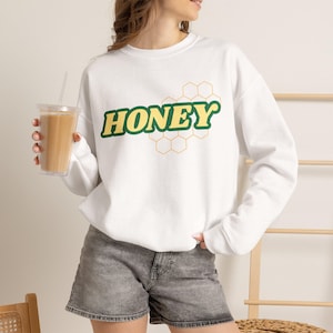 May include: A white long-sleeve crewneck sweatshirt featuring the word HONEY in yellow letters with a dark green outline, positioned above a thin orange honeycomb pattern. The sweatshirt is paired with gray denim shorts and a clear plastic cup.