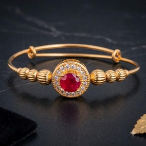 May include: A gold-toned bangle bracelet featuring a central circular setting with a round red gemstone surrounded by a halo of small clear stones. The band is accented with a series of ribbed, spherical gold-toned beads on either side of the centre.