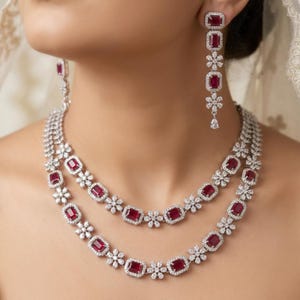 Könnte beinhalten: Ein zweireihiges Collier und passende Ohrhänger mit rechteckigen roten Edelsteinen, die von kleinen klaren Steinen umrahmt sind. Das Design umfasst abwechselnd blumenförmige Cluster aus klaren Steinen zwischen jedem roten Stein.