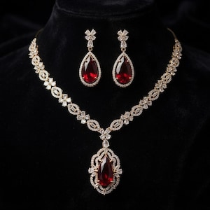 May include: A jewelry set consisting of a necklace and matching drop earrings. The necklace features a central teardrop-shaped red stone surrounded by clear crystals, with a chain of floral-shaped crystal links. The earrings mirror the design.