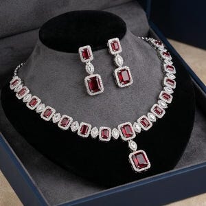 Puede incluir: Un conjunto de joyería compuesto por un collar y pendientes colgantes a juego. Cada pieza presenta piedras rectangulares de color rojo intenso facetadas, rodeadas por pequeñas piedras claras brillantes en un marco de metal plateado.