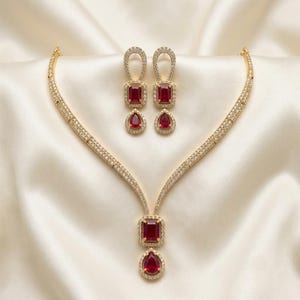 May include: A gold-toned necklace and earring set featuring clear pavé crystals and deep red stones. The necklace has a central pendant with a rectangular and a teardrop-shaped red stone. The matching earrings mirror this design with a drop style.