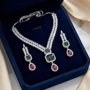 Puede incluir: Un conjunto de collar y pendientes plateados con piedras transparentes de talla marquesa en forma de hoja. El colgante y los pendientes incluyen piedras cuadradas de color verde esmeralda y azul junto a piedras rojas rubí en forma de lágrima.