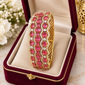 Puede incluir: Un brazalete rígido articulado en tono dorado con tres filas de piedras ovaladas de color rosa vibrante y pequeñas piedras blancas transparentes en un patrón ondulado. El brazalete tiene un diseño lateral de celosía y se exhibe en un estuche de terciopelo rojo.