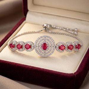 Puede incluir: Una pulsera ajustable de tono plateado con un motivo circular central de anillos concéntricos de piedras pavé transparentes que rodean una gema roja ovalada. La cadena incluye seis gemas rojas ovaladas en engastes pavé.
