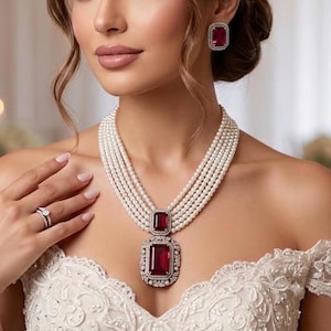 Puede incluir: Un collar de perlas de varias hebras con un gran colgante de piedra preciosa roja rectangular enmarcado por pequeñas piedras claras, un pendiente de piedra preciosa roja rectangular a juego y un anillo plateado con una piedra clara.