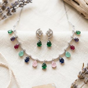 May include: A silver-toned necklace and matching earring set featuring teardrop-shaped gemstones in various colours including green, blue, pink, and purple, accented with small clear stones along the chain and earring posts.