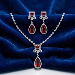 Op de afbeelding: Een sieradenset bestaande uit een ketting en twee bijpassende hangende oorbellen. Elk stuk heeft een zilverkleurige metalen zetting met heldere pavé-stenen en dieprode, peervormige en rechthoekig geslepen stenen in een symmetrisch ontwerp.