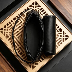 Puede incluir: Una bolsa de cuero negra con cord&oacute;n y un estuche cil&iacute;ndrico de cuero se encuentran sobre una bandeja decorativa dorada con un patr&oacute;n geom&eacute;trico. La bolsa est&aacute; abierta. El fondo es oscuro.