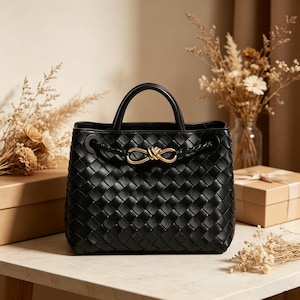 Puede incluir: Bolso de mano de cuero negro tejido con un detalle de nudo dorado. El bolso tiene una forma estructurada con un asa superior y un cierre de cord&oacute;n trenzado. El bolso est&aacute; sobre una superficie de m&aacute;rmol.