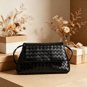 Puede incluir: Un bolso de mano negro de cuero tejido con cierre de solapa y correa para el hombro. El bolso es rectangular con esquinas redondeadas. El fondo incluye flores secas en un jarr&oacute;n y una caja de regalo.