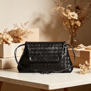 Puede incluir: Bolso de mano de cuero negro tejido con cierre de solapa y correas con cord&oacute;n. El bolso es rectangular con un patr&oacute;n tejido y una solapa plegable. El bolso est&aacute; sobre una superficie blanca con elementos decorativos en el fondo.