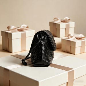 Puede incluir: Un bolso de mano negro de cuero tejido con cierre de cord&oacute;n se encuentra sobre una caja de regalo. El bolso tiene una correa para el hombro y est&aacute; rodeado de otras cajas de regalo con lazos de sat&eacute;n. El fondo es beige neutro.