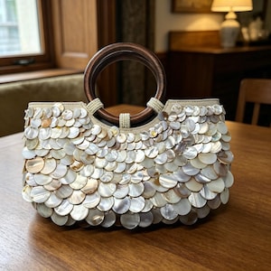 Op de afbeelding: Een crèmekleurige handtas met een houten handvat. De tas is bedekt met iriserende, cirkelvormige schelpachtige versieringen. Het handvat is een donkerbruin, cirkelvormig ontwerp. De tas staat op een houten tafel.