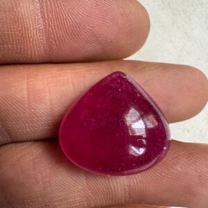 May include: A polished, teardrop-shaped gemstone in a deep, saturated magenta hue. The stone has a smooth, reflective surface, with subtle variations in color. The gem is held between fingers, showcasing its size and shape.