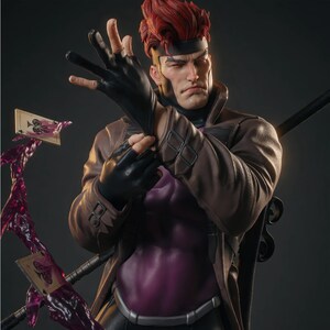 May include: A detailed collectible figurine of a character with spiky red hair, wearing a brown trench coat over a purple shirt and black gloves. The figure is posed with a black staff and two playing cards surrounded by translucent purple energy.