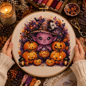 Puede incluir: Un bastidor de bordado circular con un patrón de punto de cruz de una criatura morada con sombrero de bruja, rodeada de varias calabazas naranjas y follaje otoñal morado. El diseño incluye un pequeño farol a cada lado.