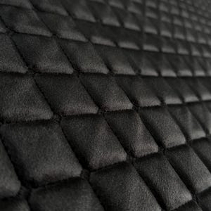 May include: A black fabric surface featuring a repeating quilted diamond pattern. The texture appears soft and slightly raised, with deep stitching lines creating a grid of individual diamond-shaped sections across the entire visible area.