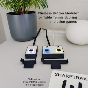 May include: Two black and white wireless button modules with illuminated squares, designed for table tennis scoring. The image text reads "Wireless Button Module for Table Tennis Scoring and other games". A white sphere and a plant in a grey pot are also visible.
