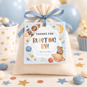 May include: A cream-colored fabric drawstring pouch tied with a blue ribbon, featuring a square tag with space-themed illustrations of a rocket, planets, stars, and a bear. The tag text reads: THANKS FOR BLASTING BY! With Love, Oliver.