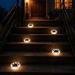 May include: Paw-shaped solar lights illuminate a wooden staircase at night. The lights are white with black paw print details, creating a warm, inviting glow. The outdoor lights are arranged on the steps.