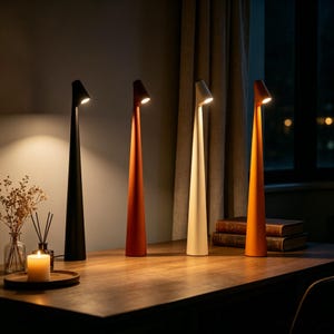 May include: Four modern cone-shaped desk lamps in black, rust, cream, and orange. Each lamp has a small, angled head that emits light. The lamps are arranged on a wooden desk, with a candle and books in the background.