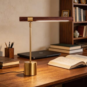 May include: A contemporary desk lamp featuring a dark wood shade and a brass base and stem. The lamp is switched on, casting light across the desk. The lamp is on a wooden desk with books and a laptop.