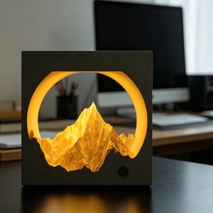 Puede incluir: Una lámpara cuadrada negra con una escena de montaña iluminada con luz amarilla cálida. La montaña es visible a través de una abertura circular, creando un efecto 3D. Un pequeño botón negro está en el lateral.