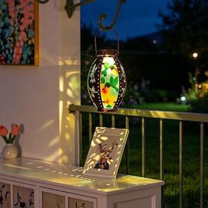 May include: A decorative outdoor lantern with a hummingbird design. The lantern is made of metal and glass, with a brown frame and colorful hummingbird images. It hangs from a metal hook on a porch, illuminated at night.