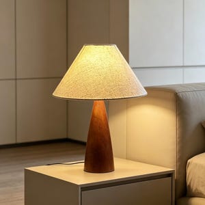 May include: A wooden table lamp with a conical, brown base and a beige fabric lampshade. The lamp is on a light-coloured bedside table. The lamp's cord is visible, and the background is softly lit.